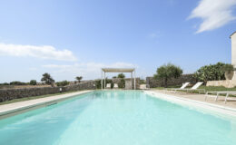 Serene outdoor swimming pool surrounded by lush greenery and a stone wall, featuring lounge chairs and a shaded pergola under a clear blue sky. Ideal for relaxation and leisure.