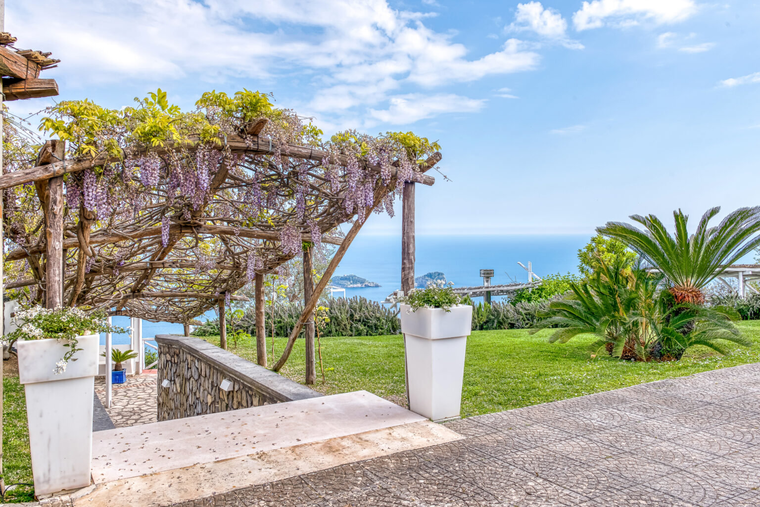 Scenic view of a garden pathway leading to the ocean, featuring a wooden pergola adorned with wisteria blossoms, lush greenery, and palm plants in a tranquil setting. | Only on SopranoVillas