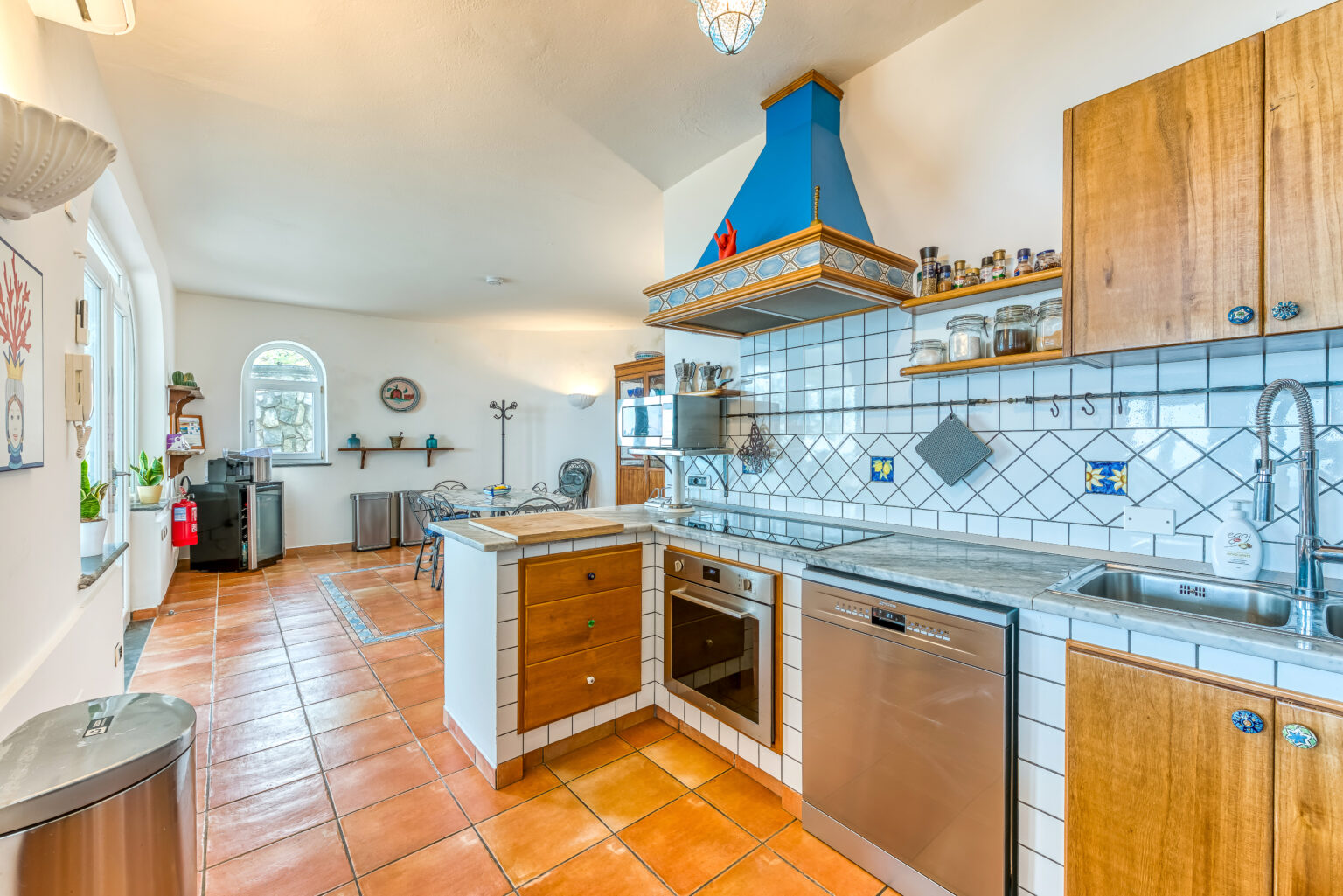 Bright and spacious kitchen featuring wooden cabinets, a blue chimney hood, and tiled countertops. The room includes stainless steel appliances, a dining area with wrought iron chairs, and ample natural light from arched windows. Terra cotta flooring enhances the warm aesthetic. | Only on SopranoVillas
