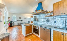 Bright and spacious kitchen featuring wooden cabinets, a blue chimney hood, and tiled countertops. The room includes stainless steel appliances, a dining area with wrought iron chairs, and ample natural light from arched windows. Terra cotta flooring enhances the warm aesthetic.