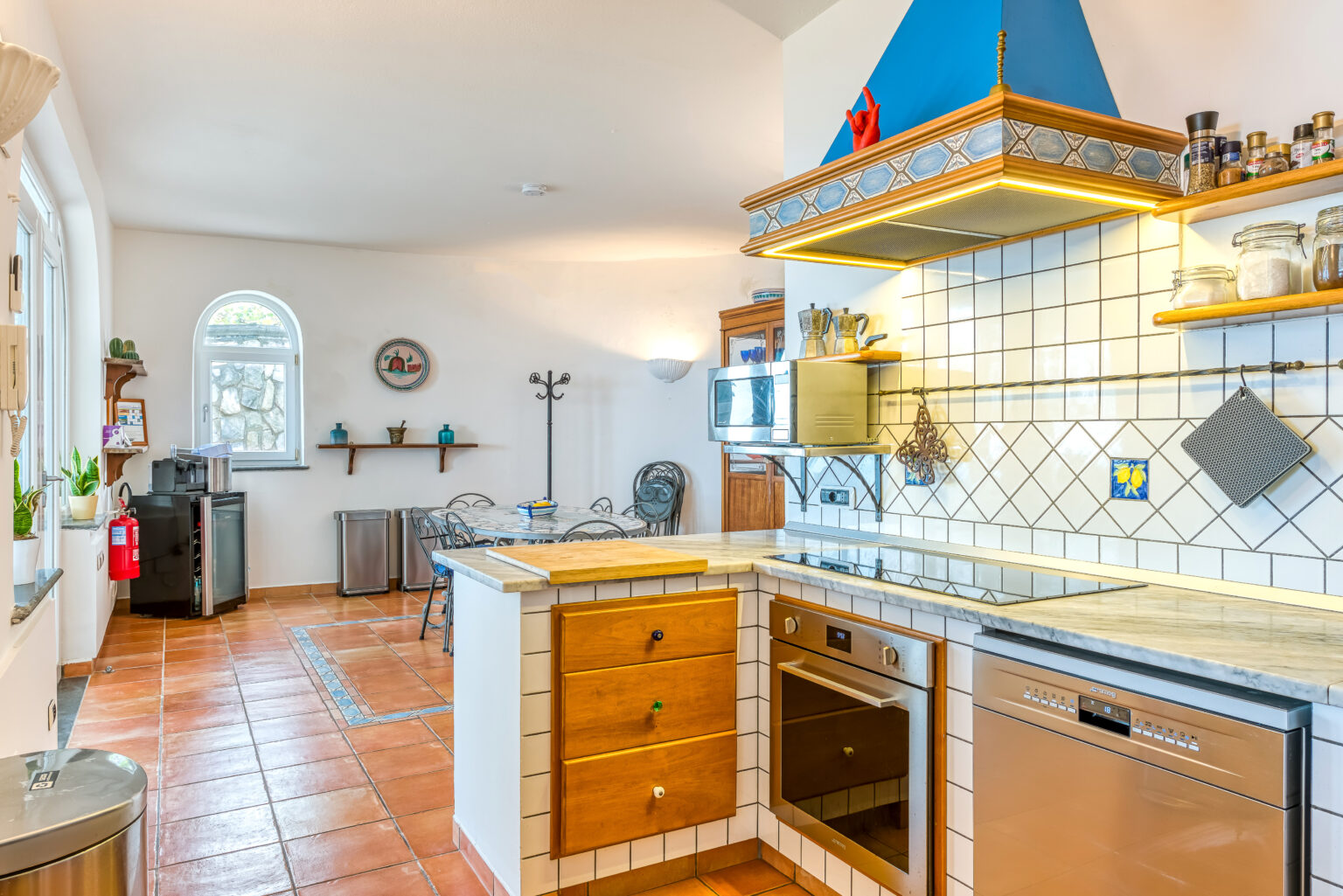 Bright and modern kitchen featuring a stylish blue hood, wooden cabinetry, and tiled countertops. The space includes an oven, dishwasher, and a cozy dining area with a marble table. Large windows provide ample natural light, enhancing the inviting atmosphere. | Only on SopranoVillas