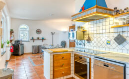 Bright and modern kitchen featuring a stylish blue hood, wooden cabinetry, and tiled countertops. The space includes an oven, dishwasher, and a cozy dining area with a marble table. Large windows provide ample natural light, enhancing the inviting atmosphere.