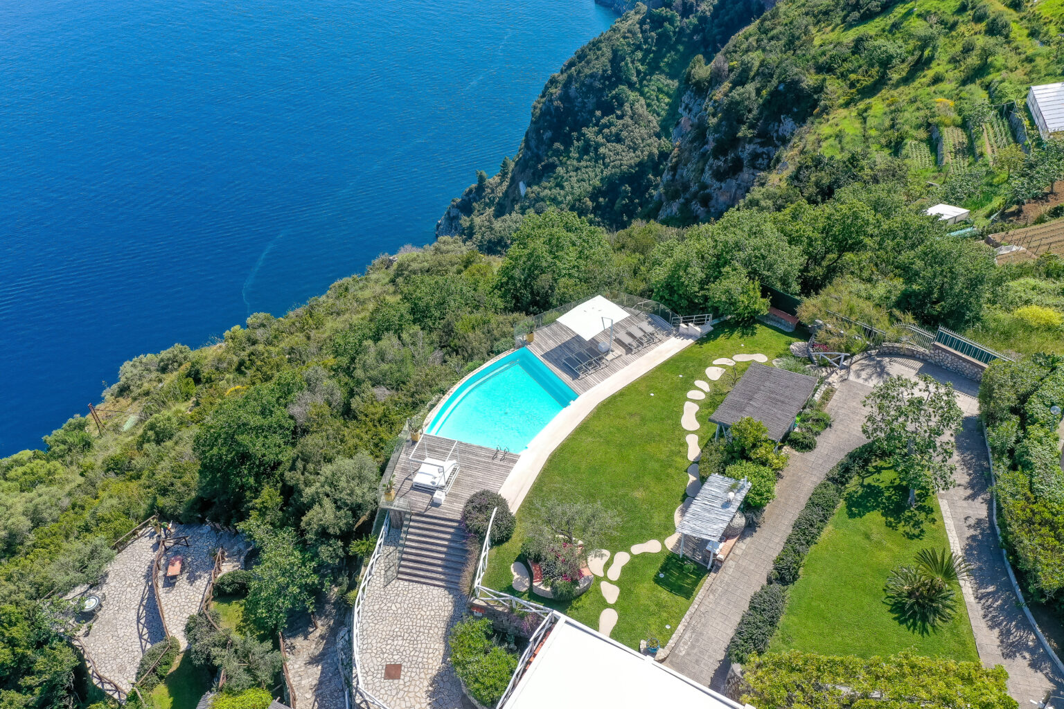 Aerial view of a luxurious villa with a swimming pool surrounded by lush greenery and terraced gardens, overlooking a stunning blue ocean. | Only on SopranoVillas