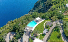 Aerial view of a luxurious villa with a swimming pool surrounded by lush greenery and terraced gardens, overlooking a stunning blue ocean.