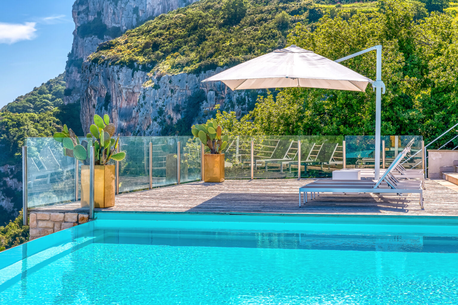 Luxury outdoor pool with a stunning mountain view, featuring a sun lounger and a large umbrella, surrounded by lush greenery and modern glass fencing. | Only on SopranoVillas