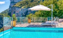 Luxury outdoor pool with a stunning mountain view, featuring a sun lounger and a large umbrella, surrounded by lush greenery and modern glass fencing.