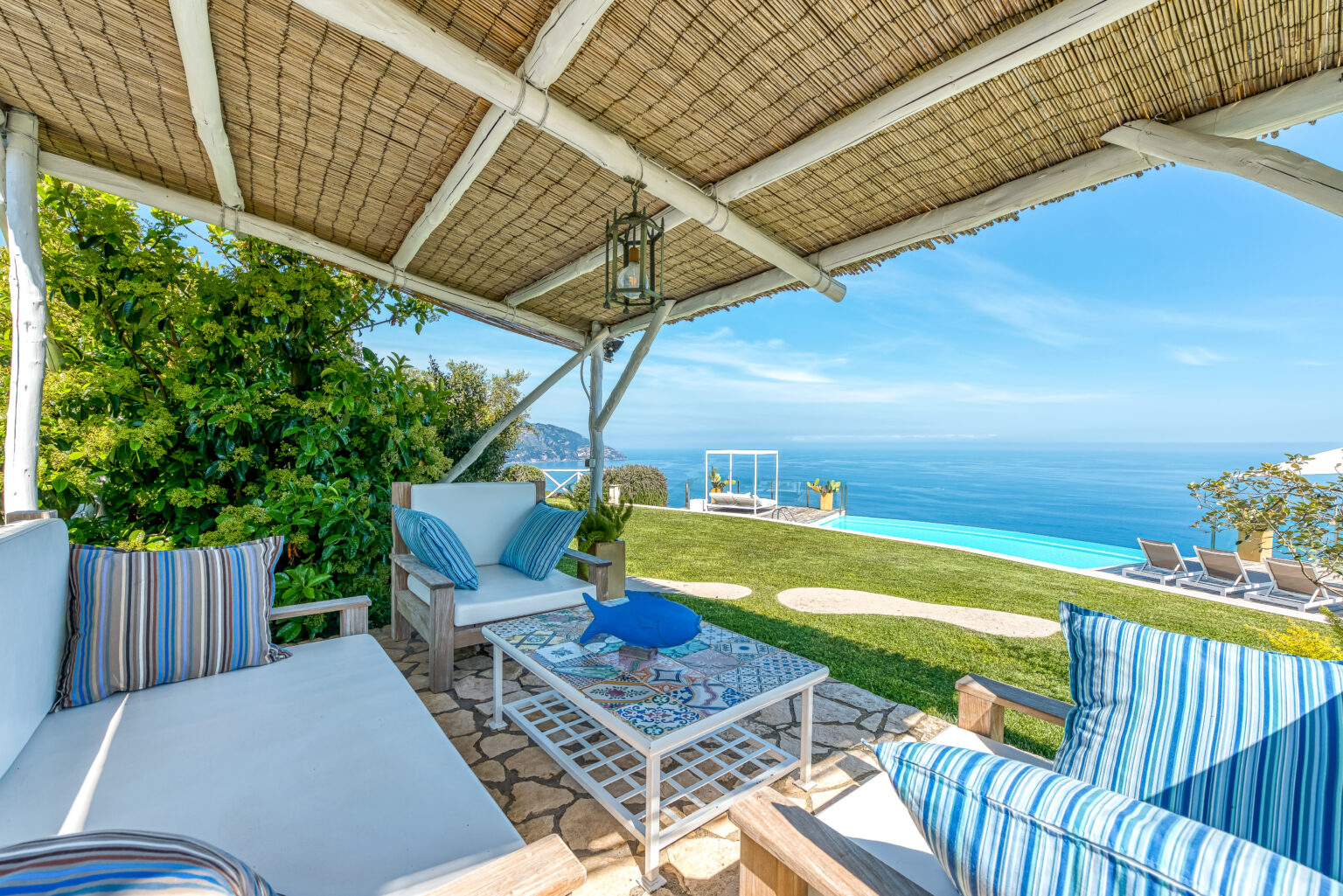 Cozy outdoor seating area with a stunning ocean view, featuring comfortable cushions and a stylish coffee table under a thatched roof. Perfect for relaxation in a luxurious villa setting. | Only on SopranoVillas