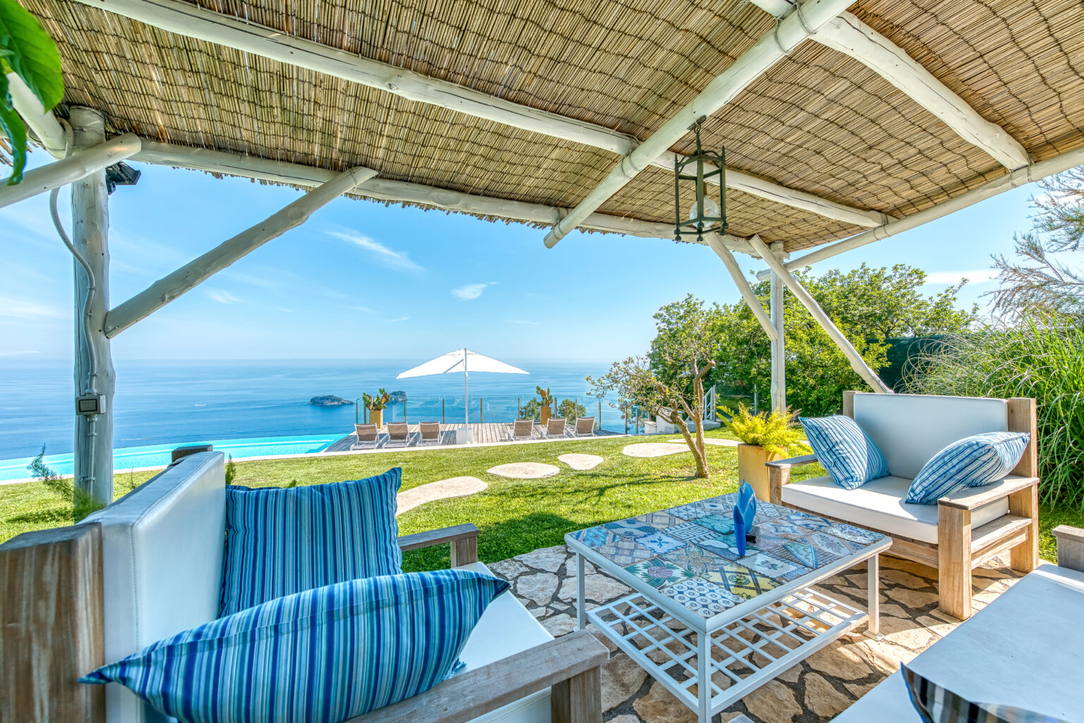 Cozy outdoor seating area with a view of the ocean, featuring a shaded pergola, comfortable furniture, and a glass coffee table, perfect for relaxation in a scenic landscape. | Only on SopranoVillas