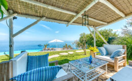 Cozy outdoor seating area with a view of the ocean, featuring a shaded pergola, comfortable furniture, and a glass coffee table, perfect for relaxation in a scenic landscape.