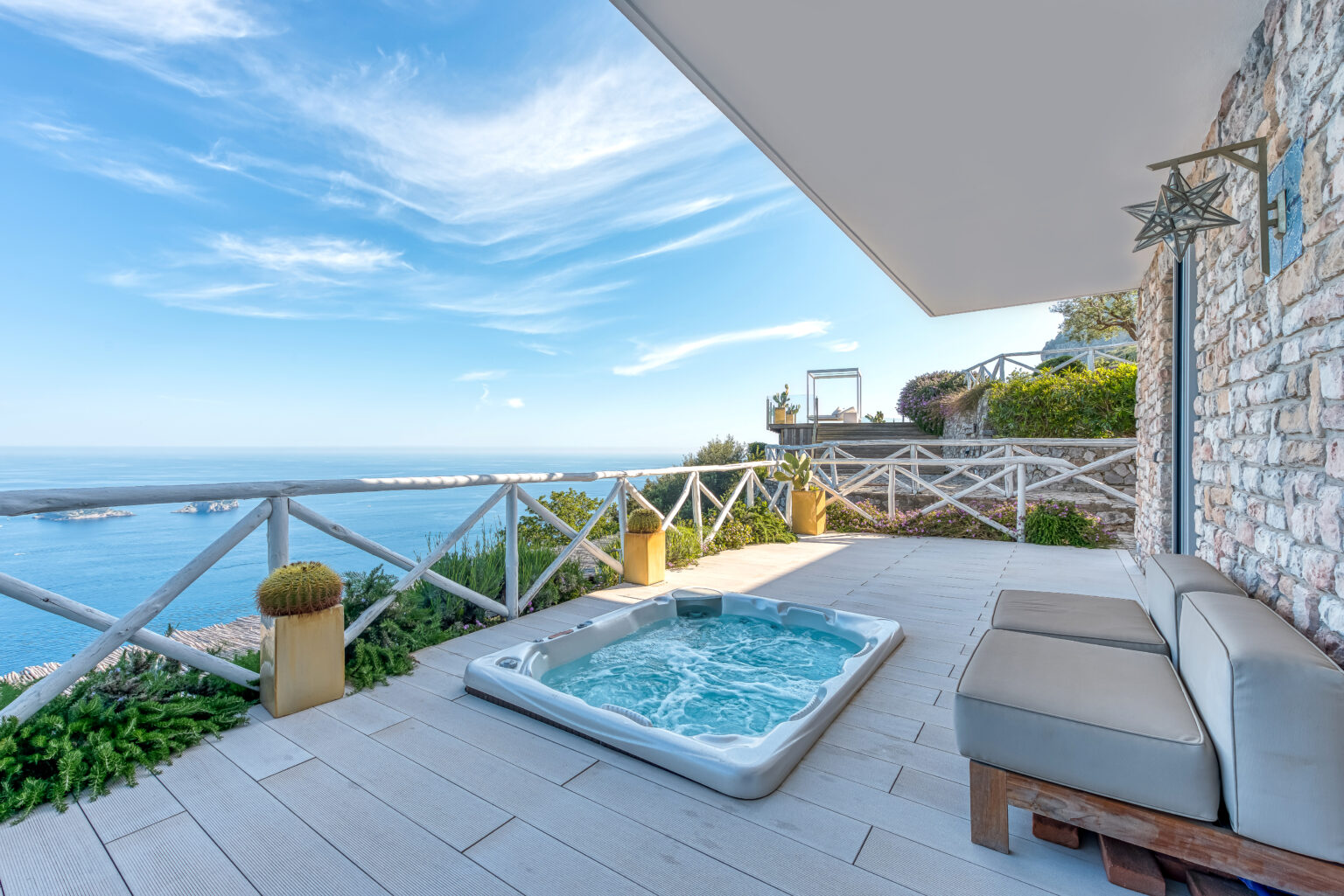 Luxurious outdoor terrace with a hot tub overlooking the ocean, surrounded by greenery and a scenic view of the sky. Perfect for relaxation and enjoying coastal scenery. | Only on SopranoVillas