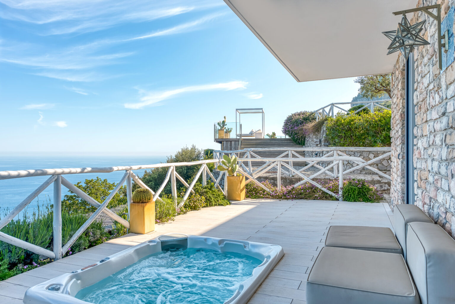 Luxurious outdoor patio with a hot tub overlooking the ocean, featuring comfortable loungers and a scenic view of the coastline under a clear blue sky. | Only on SopranoVillas