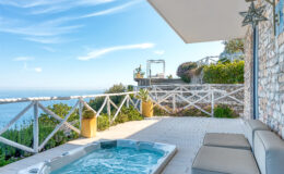 Luxurious outdoor patio with a hot tub overlooking the ocean, featuring comfortable loungers and a scenic view of the coastline under a clear blue sky.