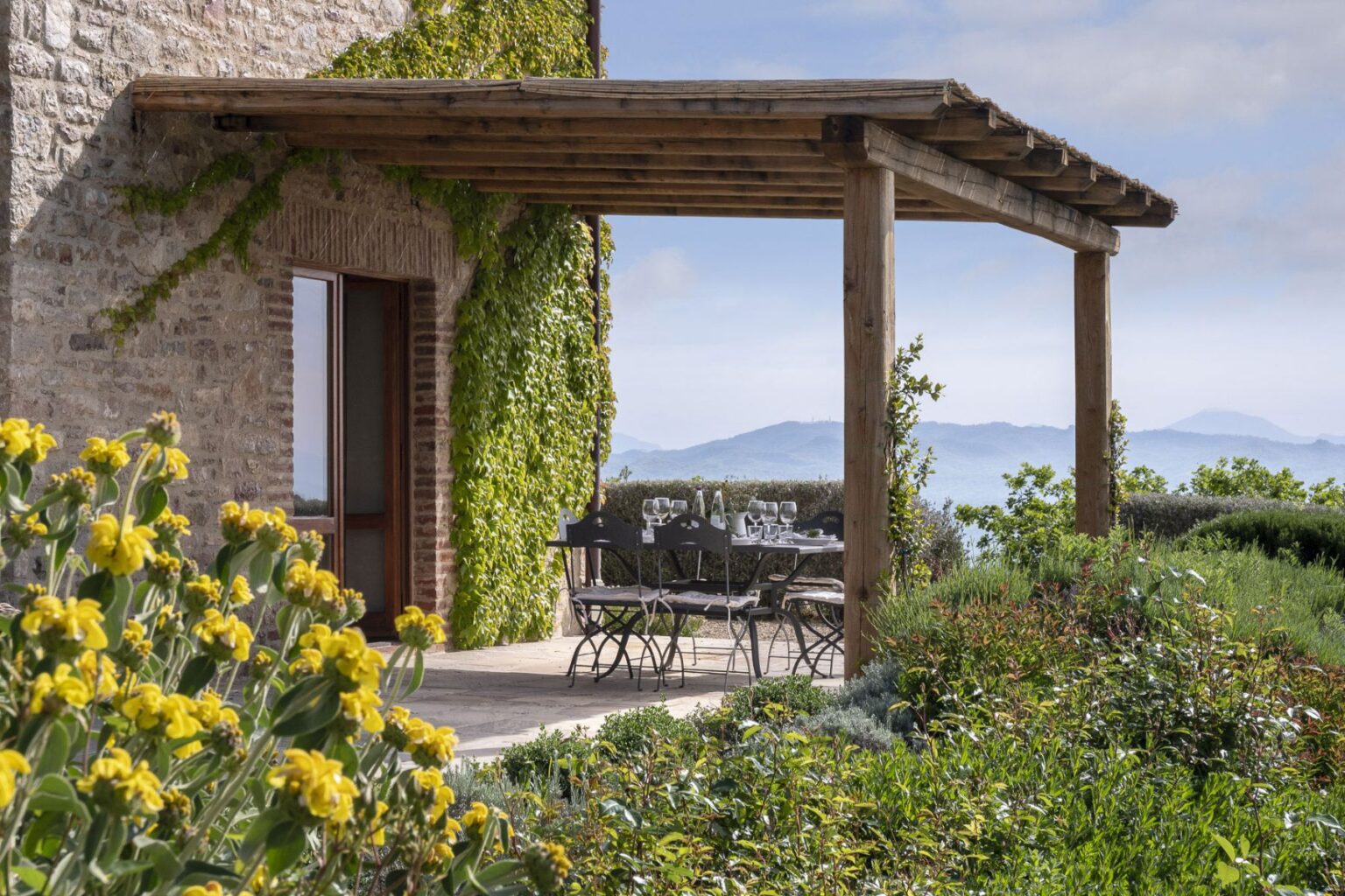 Charming outdoor dining area with a wooden pergola, featuring a table set for a meal, surrounded by vibrant flowers and lush greenery, offering scenic views of distant mountains and a clear sky. Ideal for alfresco dining in a tranquil setting. | Only on SopranoVillas