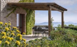 Charming outdoor dining area with a wooden pergola, featuring a table set for a meal, surrounded by vibrant flowers and lush greenery, offering scenic views of distant mountains and a clear sky. Ideal for alfresco dining in a tranquil setting.