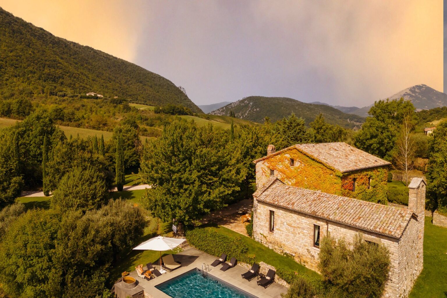 Aerial view of a rustic stone villa surrounded by lush greenery and mountains, featuring a swimming pool and lounge chairs under a sun umbrella, set in a tranquil countryside landscape. | Only on SopranoVillas