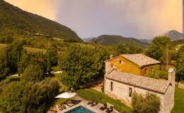 Aerial view of a rustic stone villa surrounded by lush greenery and mountains, featuring a swimming pool and lounge chairs under a sun umbrella, set in a tranquil countryside landscape.