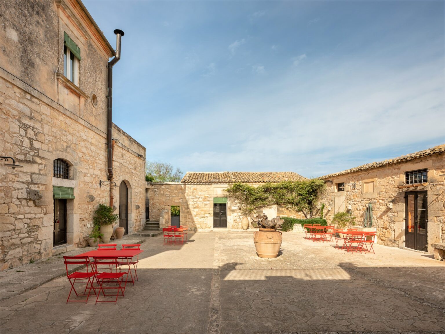 Charming courtyard featuring rustic stone buildings, vibrant red metal chairs, and a central planter, set against a clear blue sky. Ideal for outdoor dining or relaxation in a serene environment. | Only on SopranoVillas