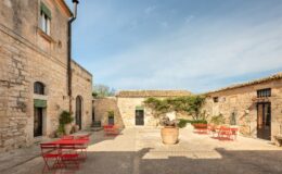 Charming courtyard featuring rustic stone buildings, vibrant red metal chairs, and a central planter, set against a clear blue sky. Ideal for outdoor dining or relaxation in a serene environment.