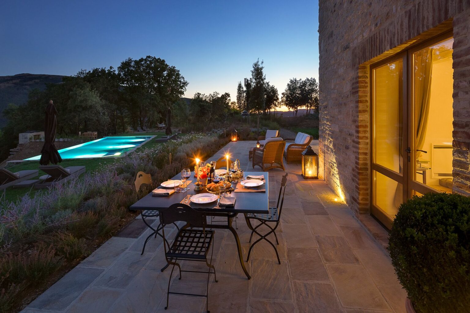 Outdoor dining setup at dusk with a beautifully arranged table, candles, and glassware, overlooking a serene pool and lush garden landscape. | Only on SopranoVillas