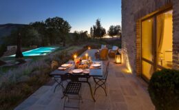 Outdoor dining setup at dusk with a beautifully arranged table, candles, and glassware, overlooking a serene pool and lush garden landscape.