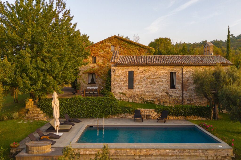 Aerial view of a rustic stone villa surrounded by lush greenery, featuring a swimming pool with lounge chairs and a hot tub, set against a serene landscape in the countryside. | Only on SopranoVillas