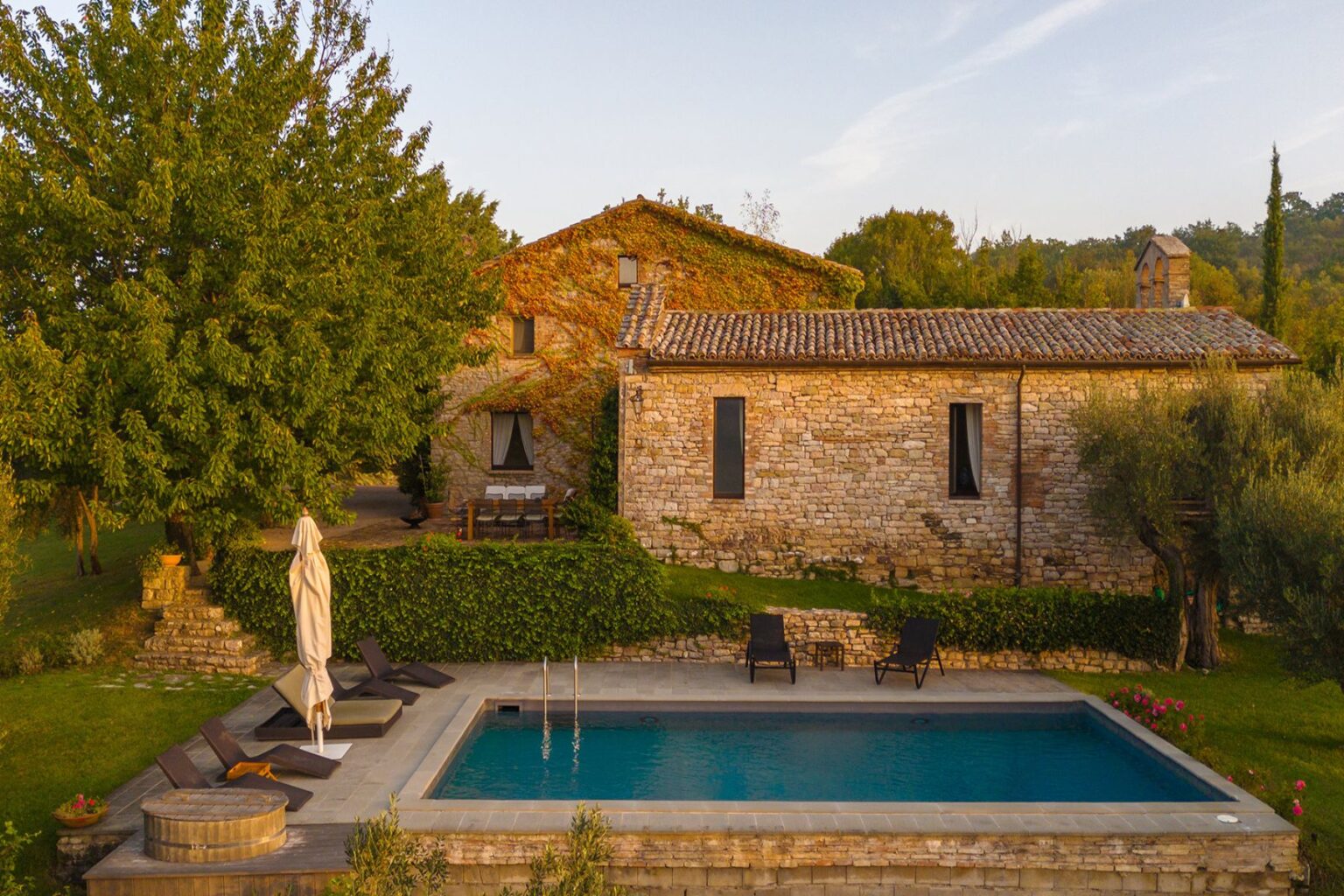 Aerial view of a rustic stone villa surrounded by lush greenery, featuring a swimming pool with lounge chairs and a hot tub, set against a serene landscape in the countryside. | Only on SopranoVillas