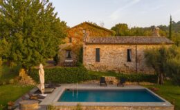 Aerial view of a rustic stone villa surrounded by lush greenery, featuring a swimming pool with lounge chairs and a hot tub, set against a serene landscape in the countryside.