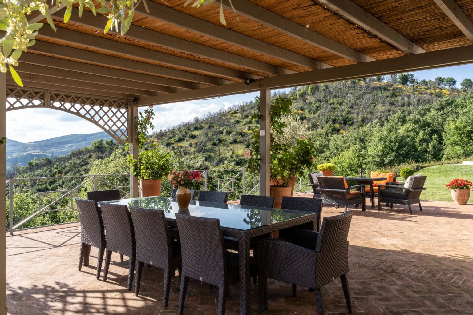 Outdoor dining area with a large glass table and wicker chairs, surrounded by lush greenery and potted plants, offering scenic views of rolling hills and a relaxing atmosphere. | Only on SopranoVillas