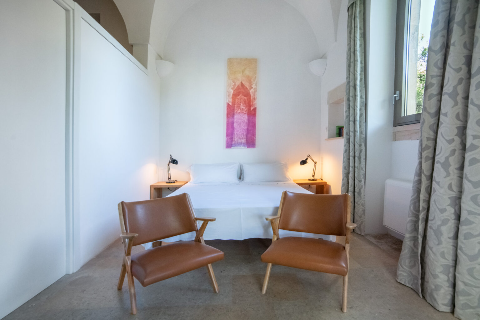 Modern bedroom interior featuring a double bed with white linens, two stylish wooden chairs, and a colorful wall art piece. Soft lighting from bedside lamps enhances the cozy atmosphere, with large windows allowing natural light to fill the space. | Only on SopranoVillas