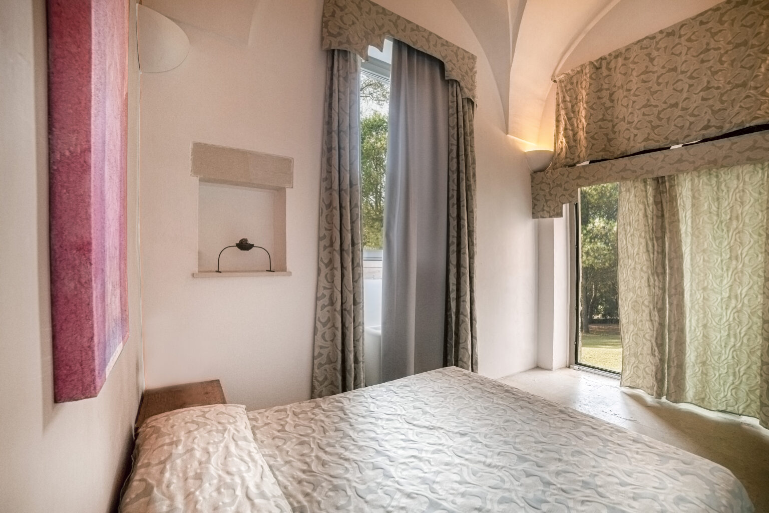 Cozy bedroom featuring a textured white bedspread, large windows with elegant curtains, and a minimalist design. The room is adorned with soft lighting and a decorative wall niche, providing a serene and inviting atmosphere. Ideal for relaxation and comfort. | Only on SopranoVillas