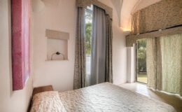 Cozy bedroom featuring a textured white bedspread, large windows with elegant curtains, and a minimalist design. The room is adorned with soft lighting and a decorative wall niche, providing a serene and inviting atmosphere. Ideal for relaxation and comfort.