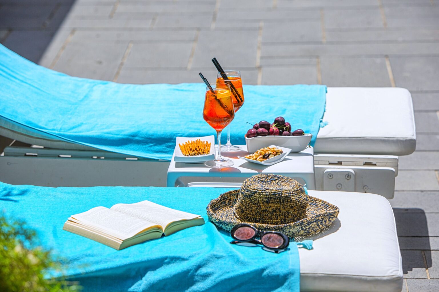 A sun lounger with a turquoise towel, featuring a book, sunglasses, a straw hat, and a plate of snacks beside two cocktails garnished with orange slices, set against a bright outdoor backdrop. | Only on SopranoVillas