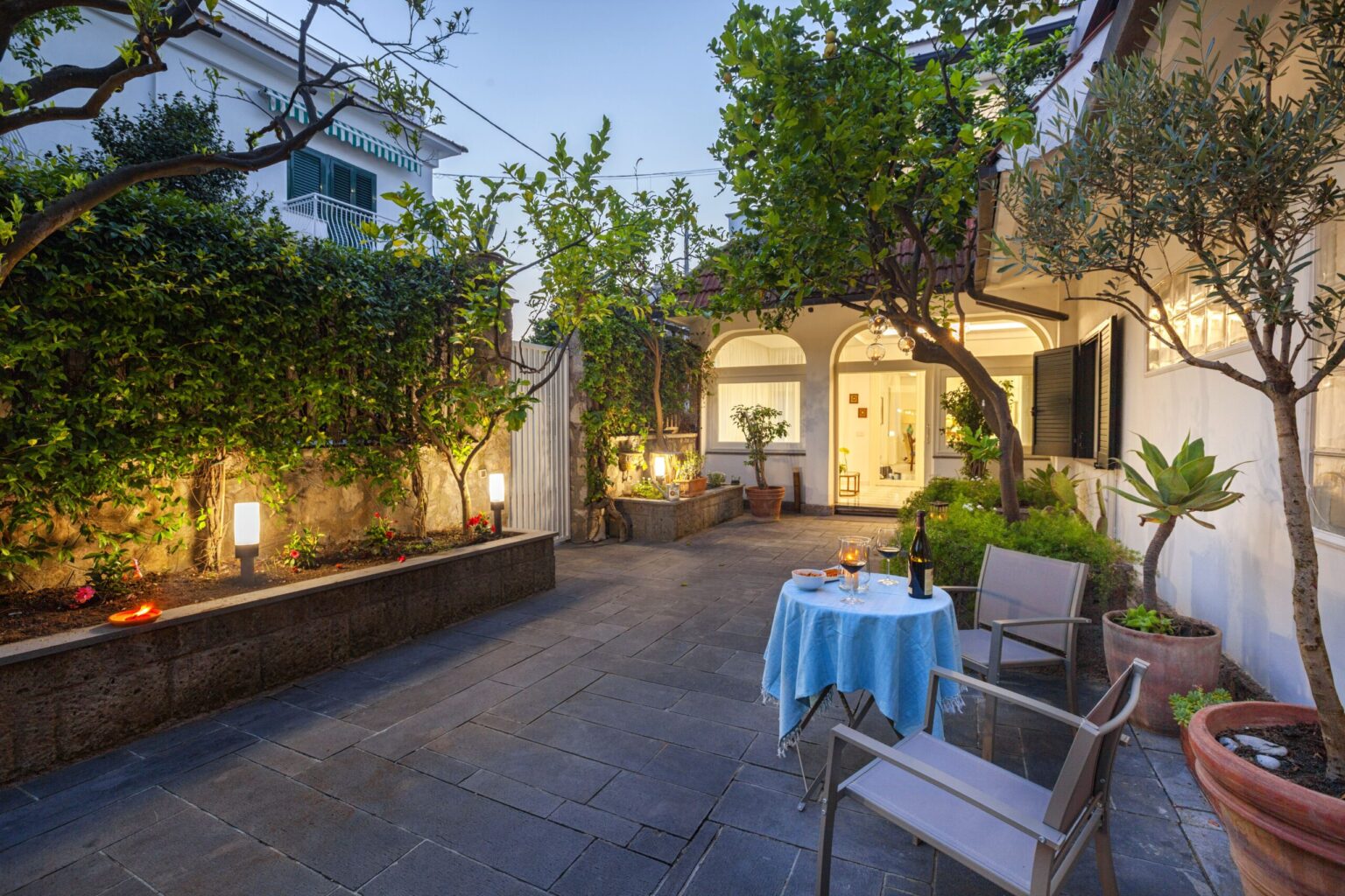 Cozy outdoor patio with a blue tablecloth, surrounded by lush greenery, potted plants, and warm lighting, ideal for relaxation and dining. | Only on SopranoVillas