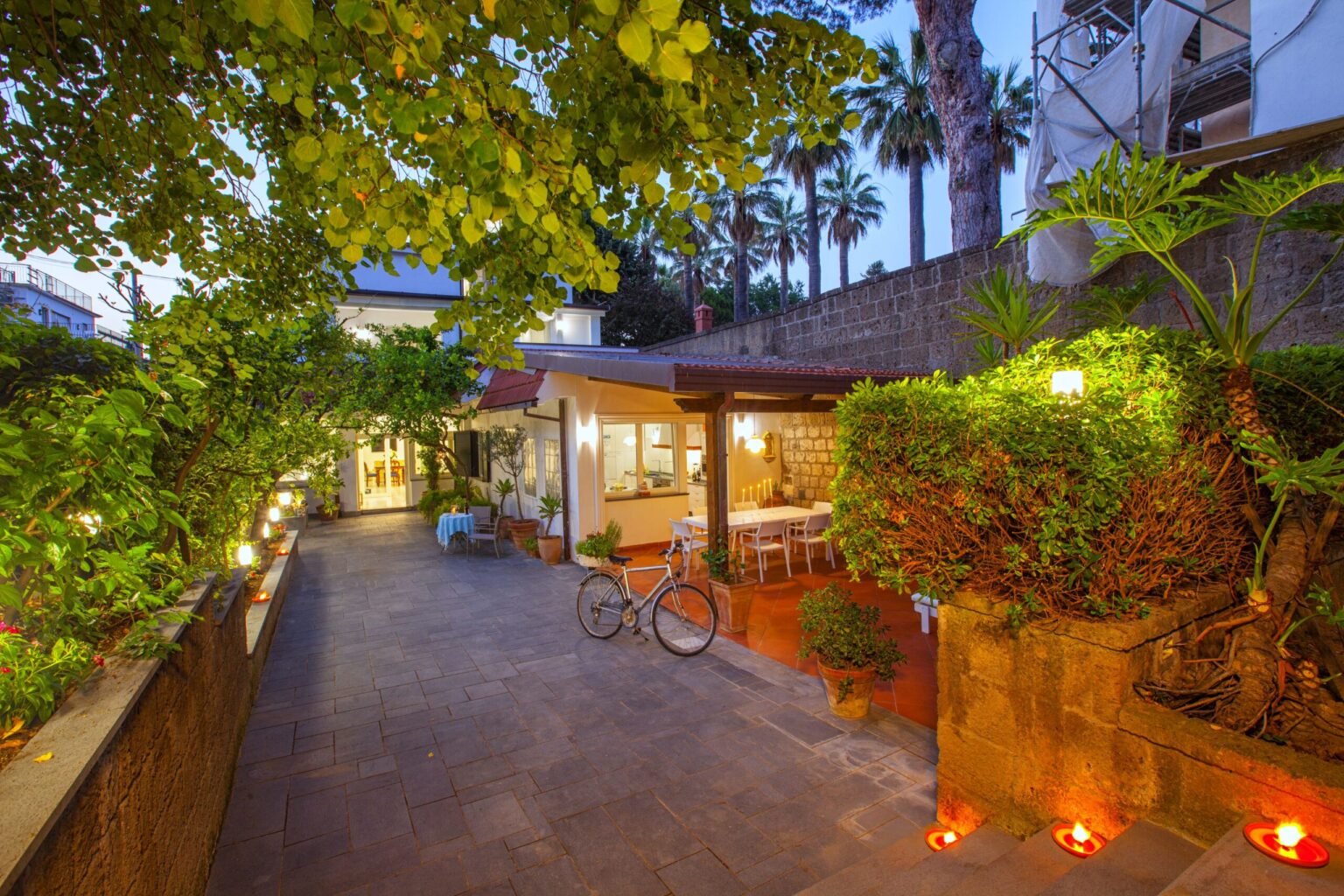 Modern home exterior with a landscaped pathway, featuring greenery and palm trees, and a cozy outdoor seating area illuminated at dusk. Ideal for showcasing contemporary architecture and outdoor living spaces. | Only on SopranoVillas
