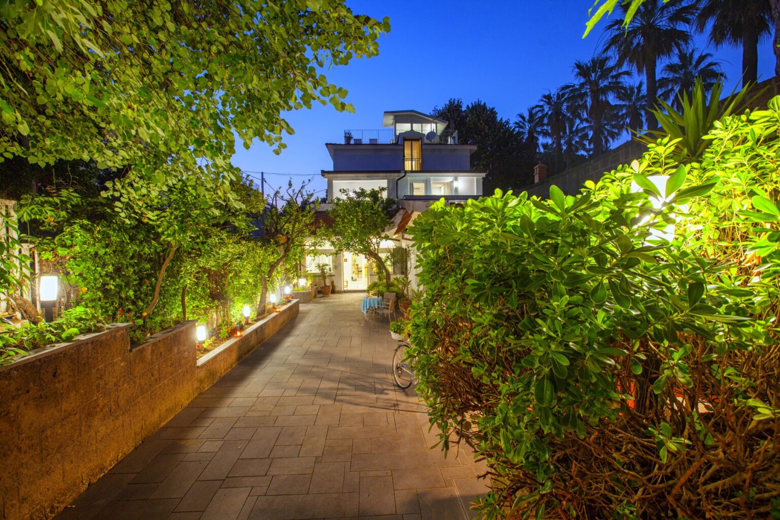 Beautiful garden pathway leading to a modern house illuminated by soft lights, surrounded by lush greenery and palm trees at dusk. Ideal for showcasing outdoor living spaces and tranquil home environments. | Only on SopranoVillas