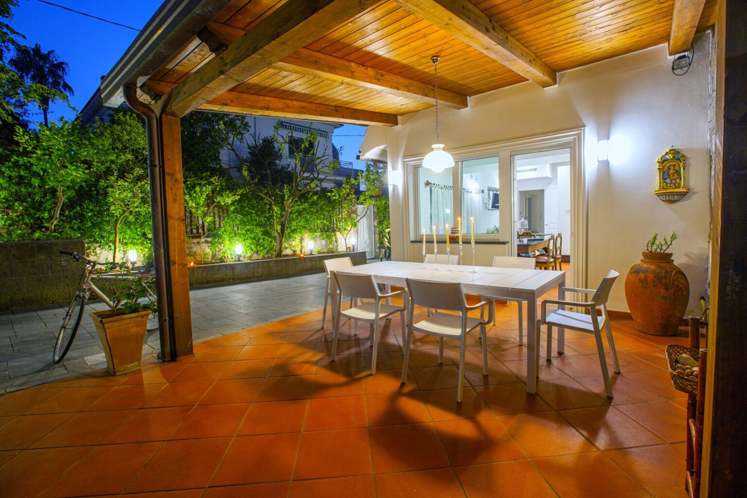 Modern outdoor dining area with a white table and chairs, surrounded by lush greenery and soft lighting, creating a cozy ambiance for evening gatherings. | Only on SopranoVillas