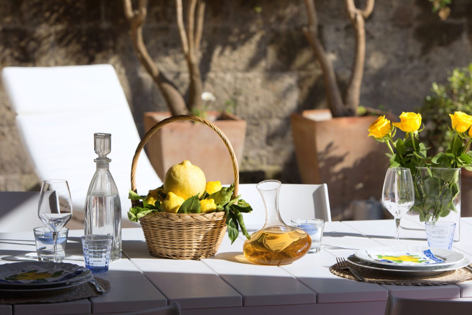 A beautifully set outdoor dining table featuring a basket of fresh lemons, a decorative yellow flower arrangement, and elegant glassware, surrounded by terracotta pots and a serene garden backdrop. | Only on SopranoVillas