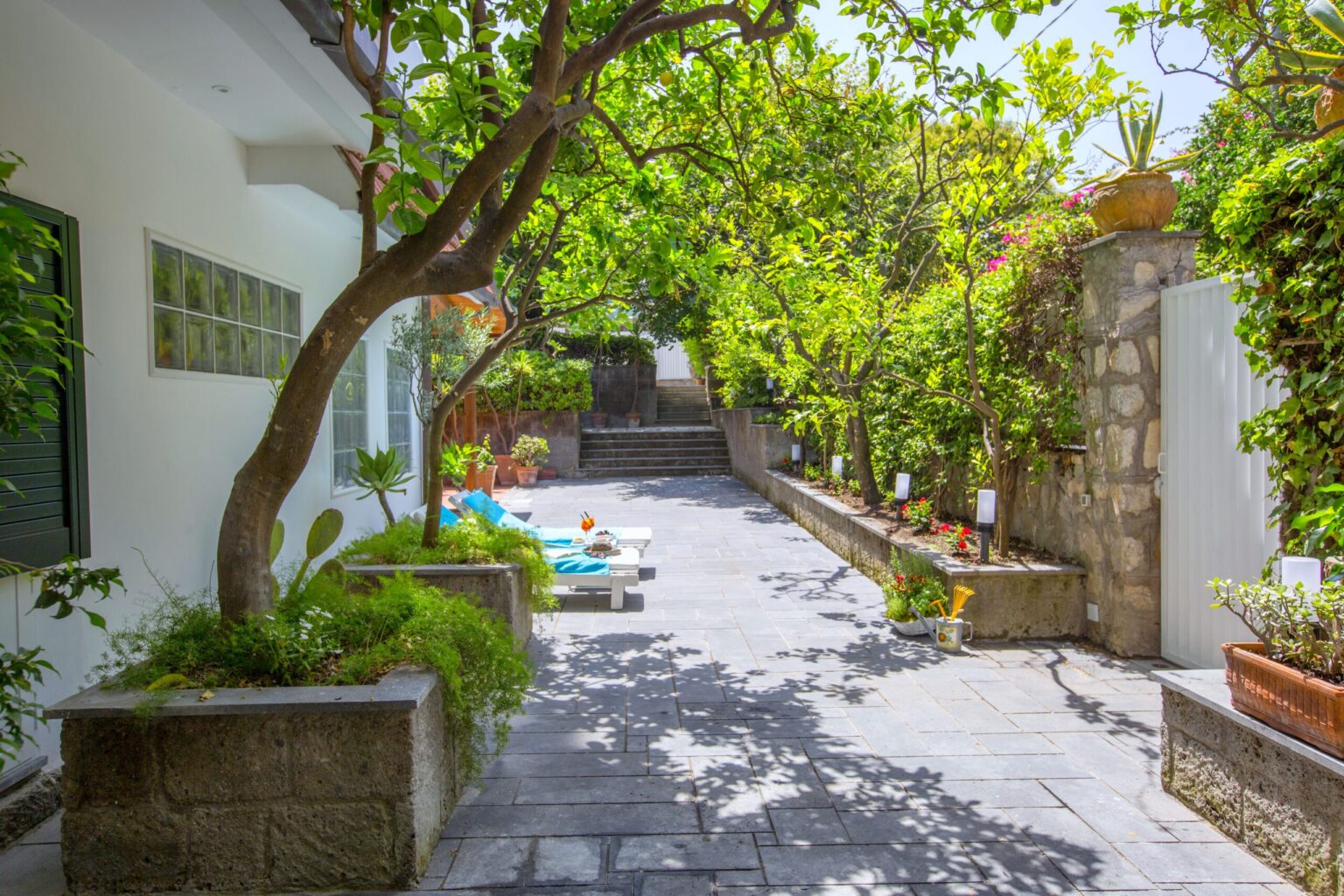Lush garden pathway leading to a serene outdoor space, surrounded by vibrant greenery and decorative plants, with a stone wall and bright sunlight illuminating the area. | Only on SopranoVillas