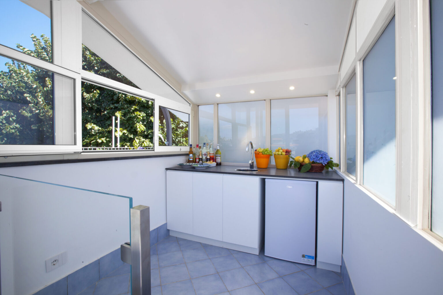 Bright and modern kitchen with large windows, featuring a countertop, sink, and refrigerator, surrounded by fresh fruits and vegetables. Ideal for cooking and entertaining. | Only on SopranoVillas