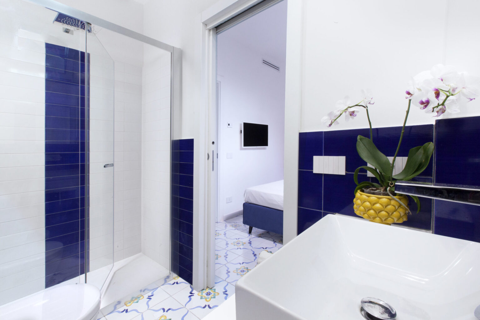 Modern bathroom featuring a walk-in shower with glass doors, blue tiled accents, and a stylish sink. A vibrant yellow planter with an orchid adds a touch of color, while the open doorway leads to a cozy bedroom. | Only on SopranoVillas