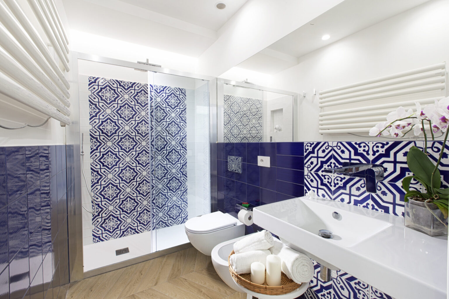 Modern bathroom featuring elegant blue and white patterned tiles, a glass shower enclosure, a sleek white sink, and a stylish towel rack. The decor includes a basket of rolled towels and a potted orchid, creating a fresh and inviting atmosphere. | Only on SopranoVillas