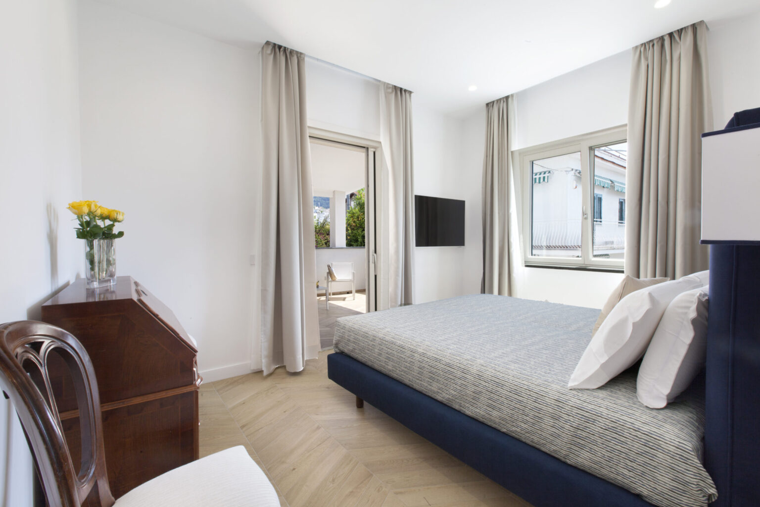 Modern bedroom featuring a blue upholstered bed with striped bedding, a wooden desk, and a vase of yellow flowers. Natural light streams in through large windows with beige curtains, revealing a view of an outdoor seating area. A wall-mounted TV adds to the contemporary decor. | Only on SopranoVillas
