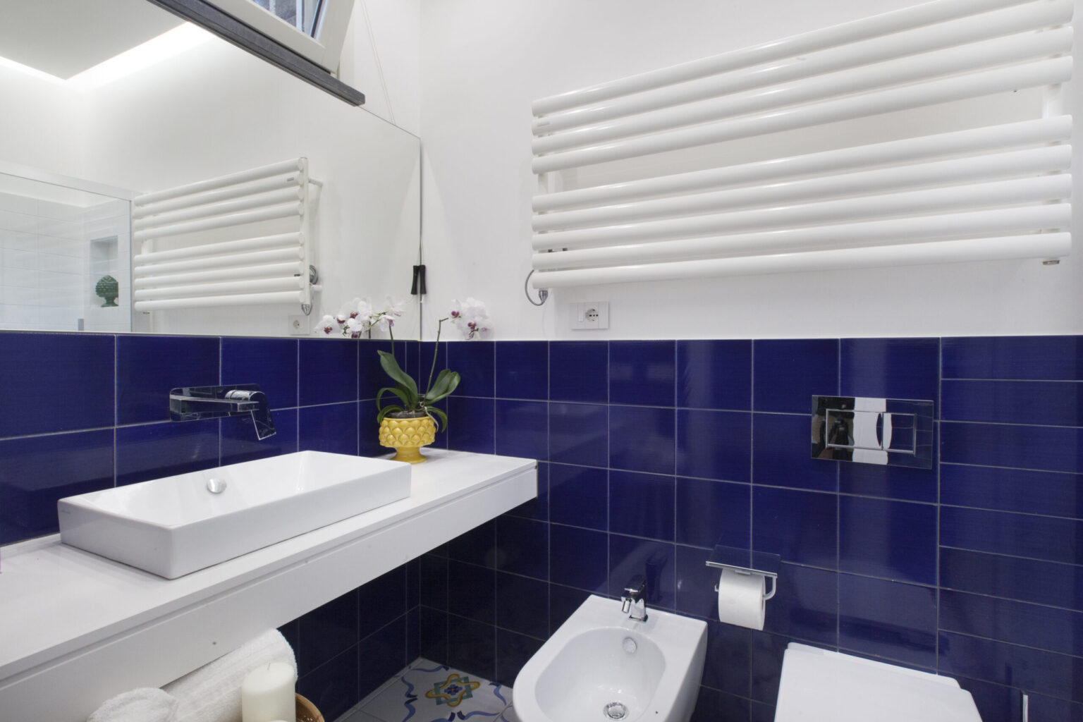 Modern bathroom design featuring a sleek white sink on a minimalist countertop, vibrant blue tiled walls, a decorative yellow plant, and a towel warmer. | Only on SopranoVillas