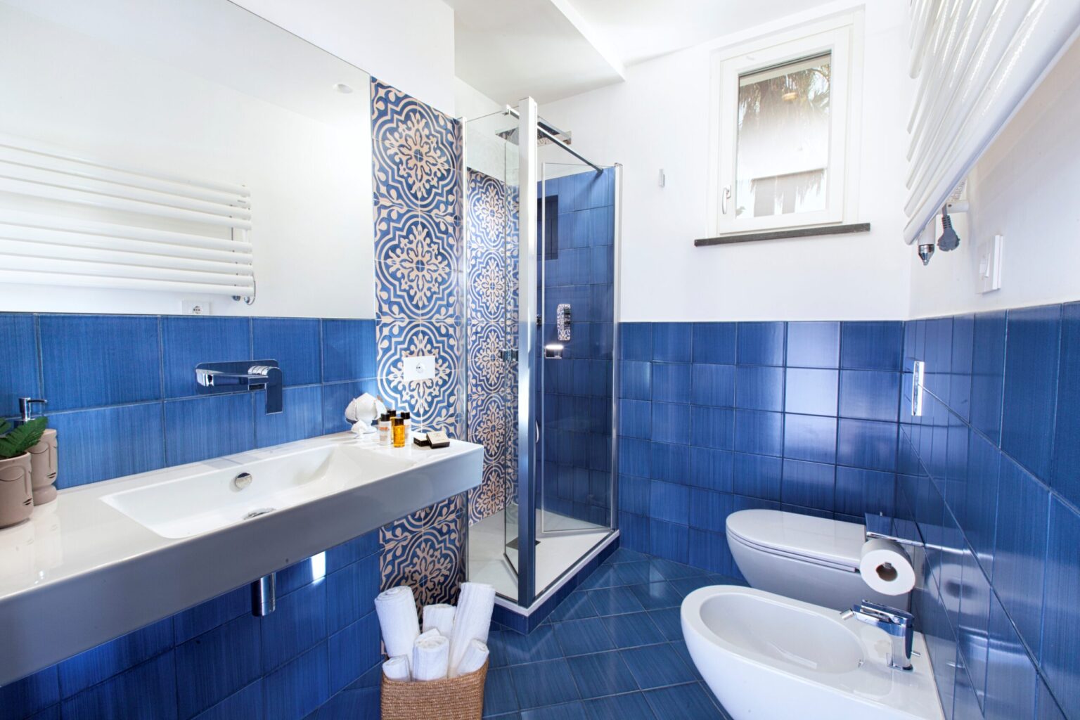 Modern bathroom featuring blue tiled walls, a glass shower enclosure, a sleek sink with toiletries, and neatly arranged towels. Ideal for showcasing contemporary bathroom design and decor. | Only on SopranoVillas