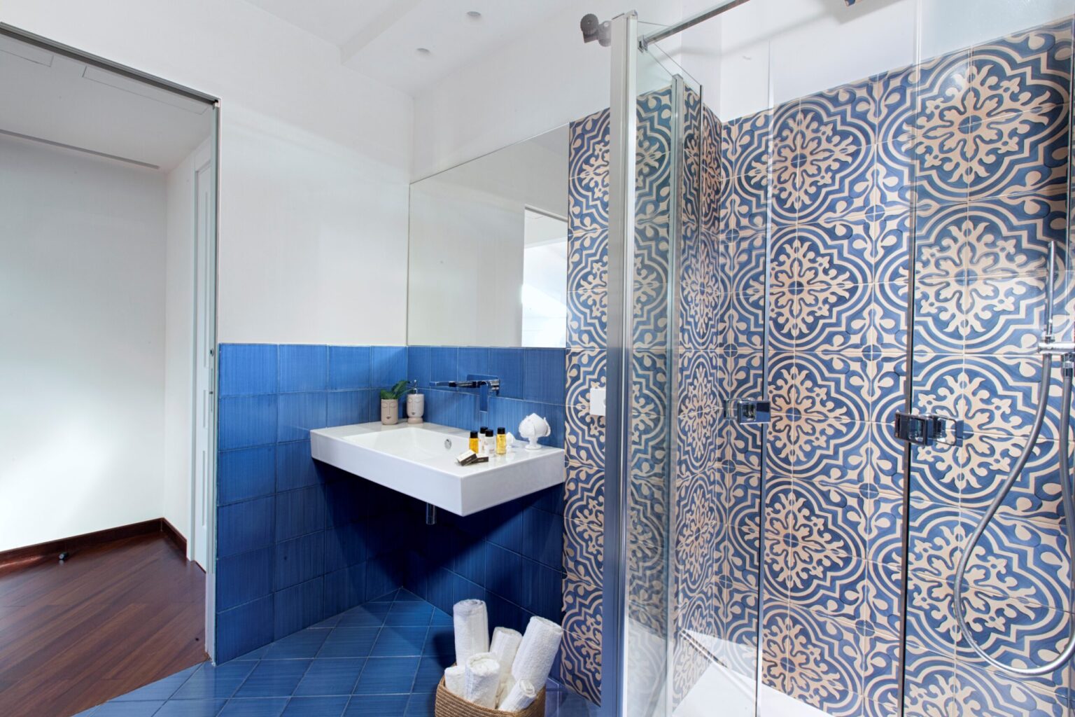 Modern bathroom featuring a glass shower enclosure, decorative blue and patterned tiles, a sleek white sink, and wooden flooring. The design combines functionality with contemporary aesthetics, creating a stylish and inviting space. | Only on SopranoVillas
