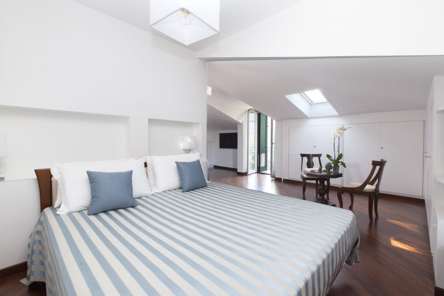 Modern bedroom featuring a striped bedspread, two blue decorative pillows, and a bright skylight. The room includes a stylish seating area with a table and chairs, complemented by wooden flooring and minimalist decor. Ideal for a relaxing stay. | Only on SopranoVillas