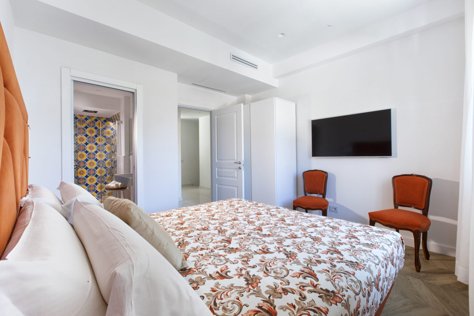 Bright and modern bedroom featuring a floral-patterned bedspread, two stylish orange chairs, and a wall-mounted television. The room has a clean, minimalist design with white walls and natural light. | Only on SopranoVillas