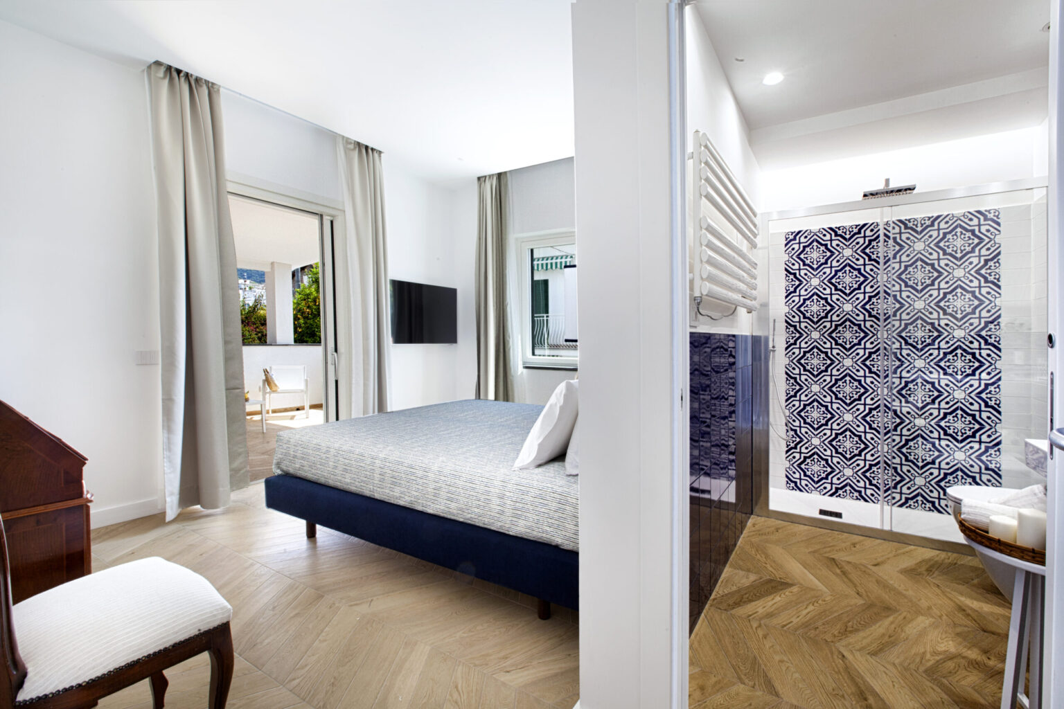 Modern bedroom with a blue bed, wooden flooring, and an adjoining bathroom featuring a stylish shower with patterned tiles. The room is well-lit, with large windows and elegant curtains, creating a spacious and inviting atmosphere. | Only on SopranoVillas