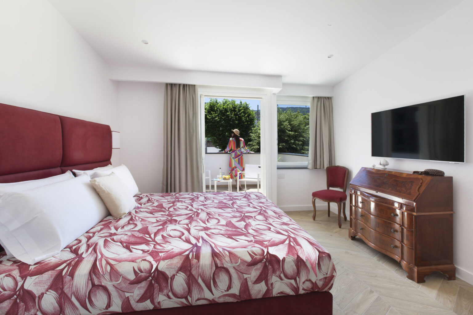 Modern hotel room featuring a red floral-patterned bedspread, white linens, and a stylish wooden dresser. A large window with curtains allows natural light, revealing a view of greenery outside. A woman in a colorful dress stands on the balcony, enhancing the inviting atmosphere. The room includes a flat-screen TV and a red chair, creating a comfortable and elegant space for relaxation. | Only on SopranoVillas