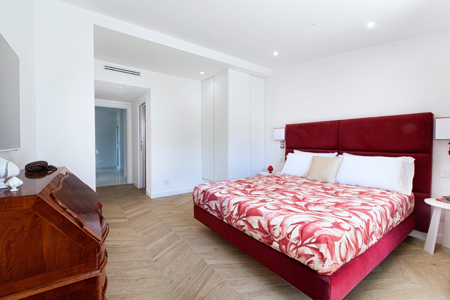 Modern bedroom featuring a red floral-patterned bedspread, minimalist decor, and ample natural light. Includes a wooden desk and stylish white walls, creating a relaxing atmosphere. Perfect for a cozy getaway or home design inspiration. | Only on SopranoVillas